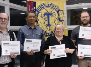 Quatre personnes souriantes tenant chacune un certificat devant une bannière arborant le logo des Toastmasters.