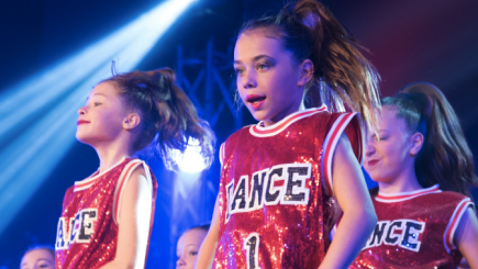 Trois jeunes filles sur scène à l'occasion d'un spectacle de danse, toutes vêtues d'une camisole sportive à paillettes rouges portant l'inscription DANCE.