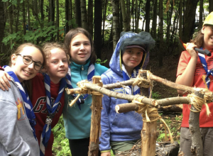 Jeunes filles devant une construction faite de bouts de bois et de cordages, avec une forêt à l'arrière.